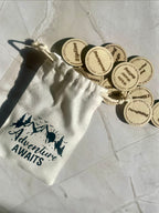 White drawstring bag with 'Adventure Awaits' text and wooden tokens on a light stone floor.