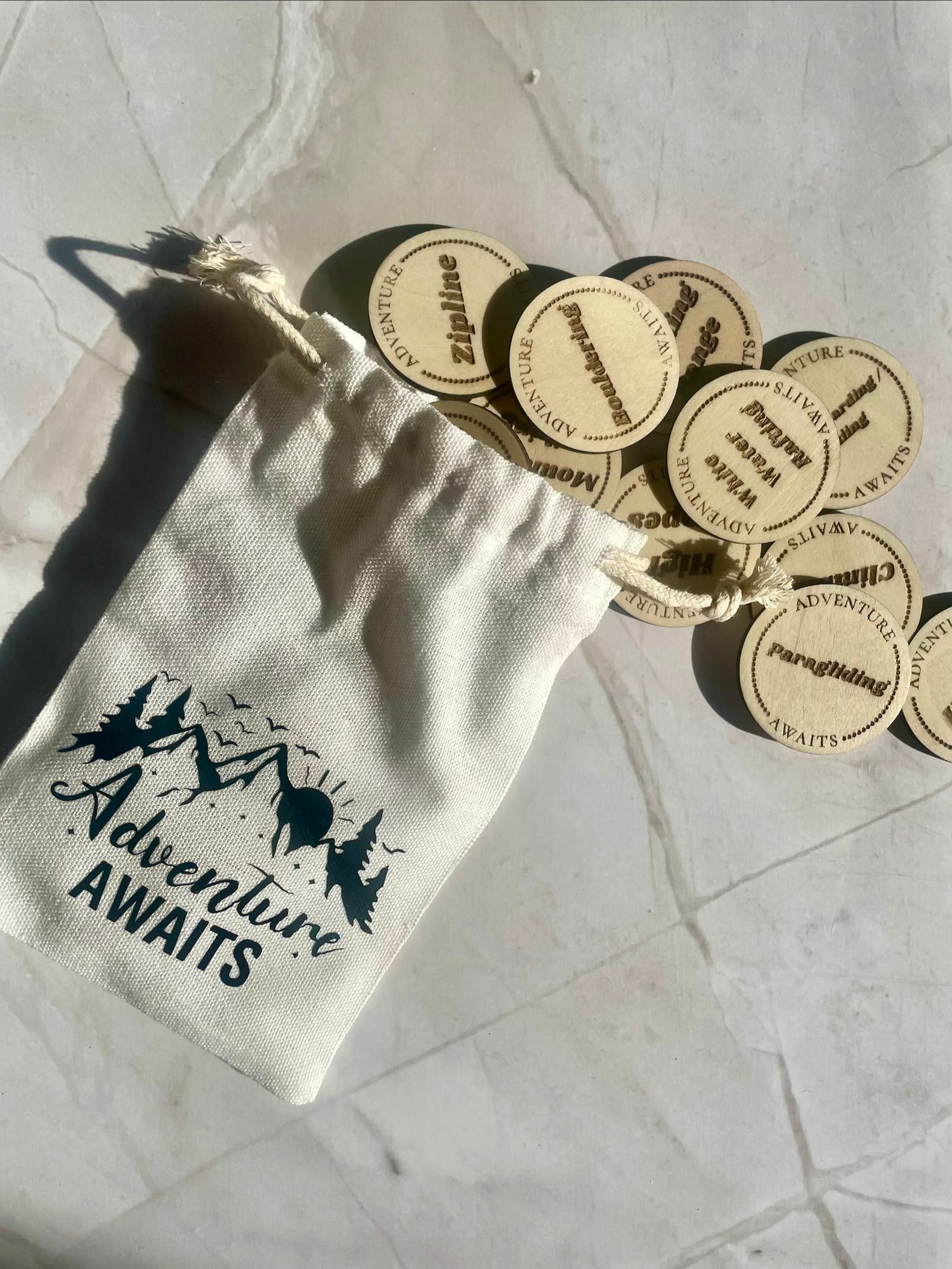 White drawstring bag with 'Adventure Awaits' text and wooden tokens on a light stone floor.