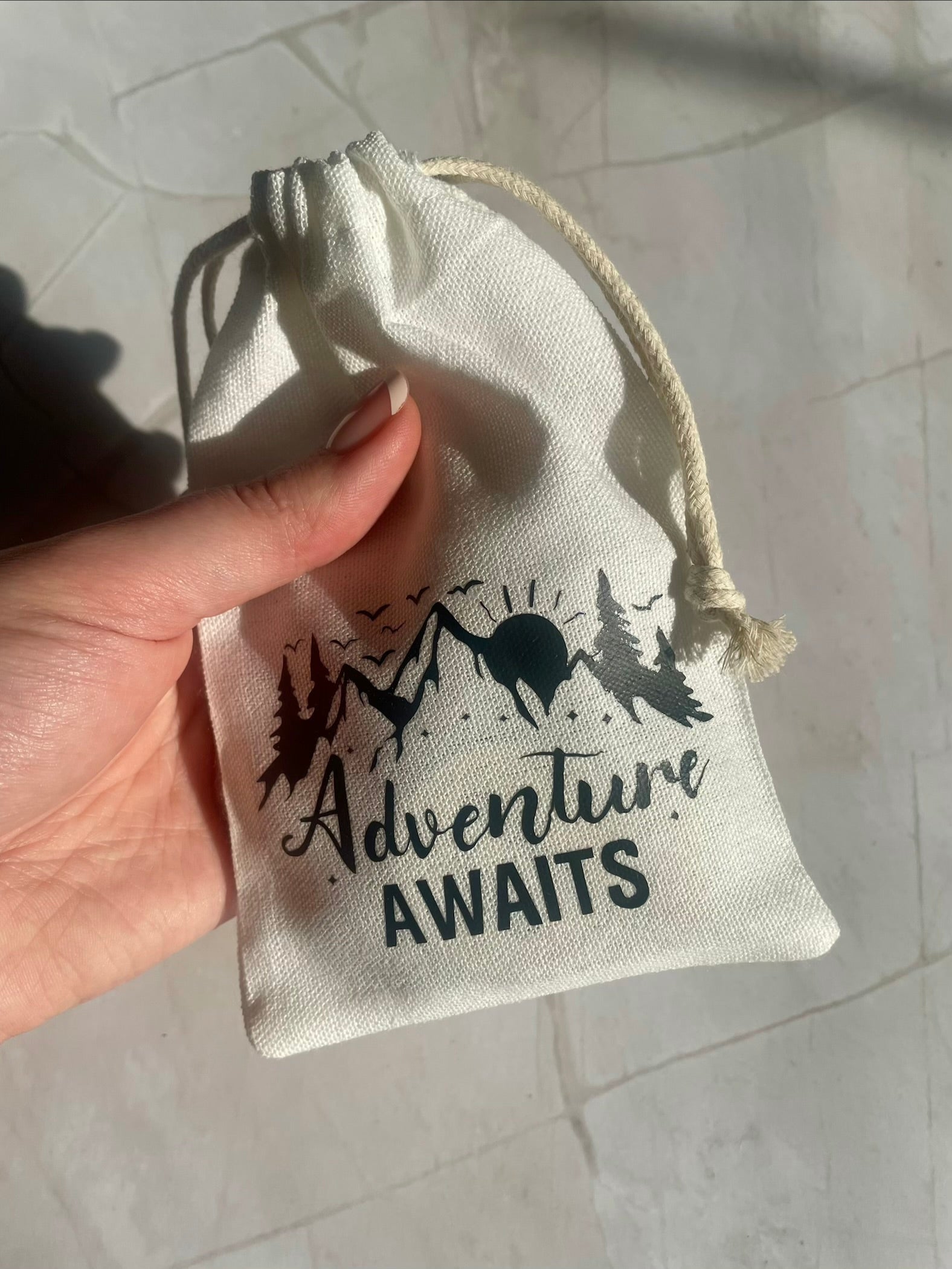 Hand holding a small drawstring bag with 'Adventure Awaits' text and mountain design.