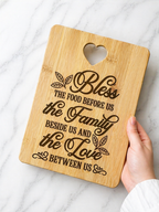 Hand holding bamboo chopping board engraved with “Bless the food before us the family beside us and the love between us” quote on marble background