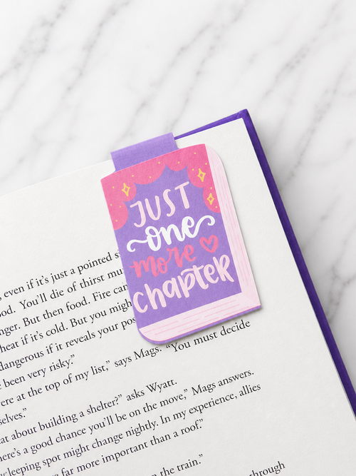 Magnetic bookmark clipped onto the top of an open book page, featuring a purple and pink “Just one more chapter” design, demonstrating how it holds pages securely