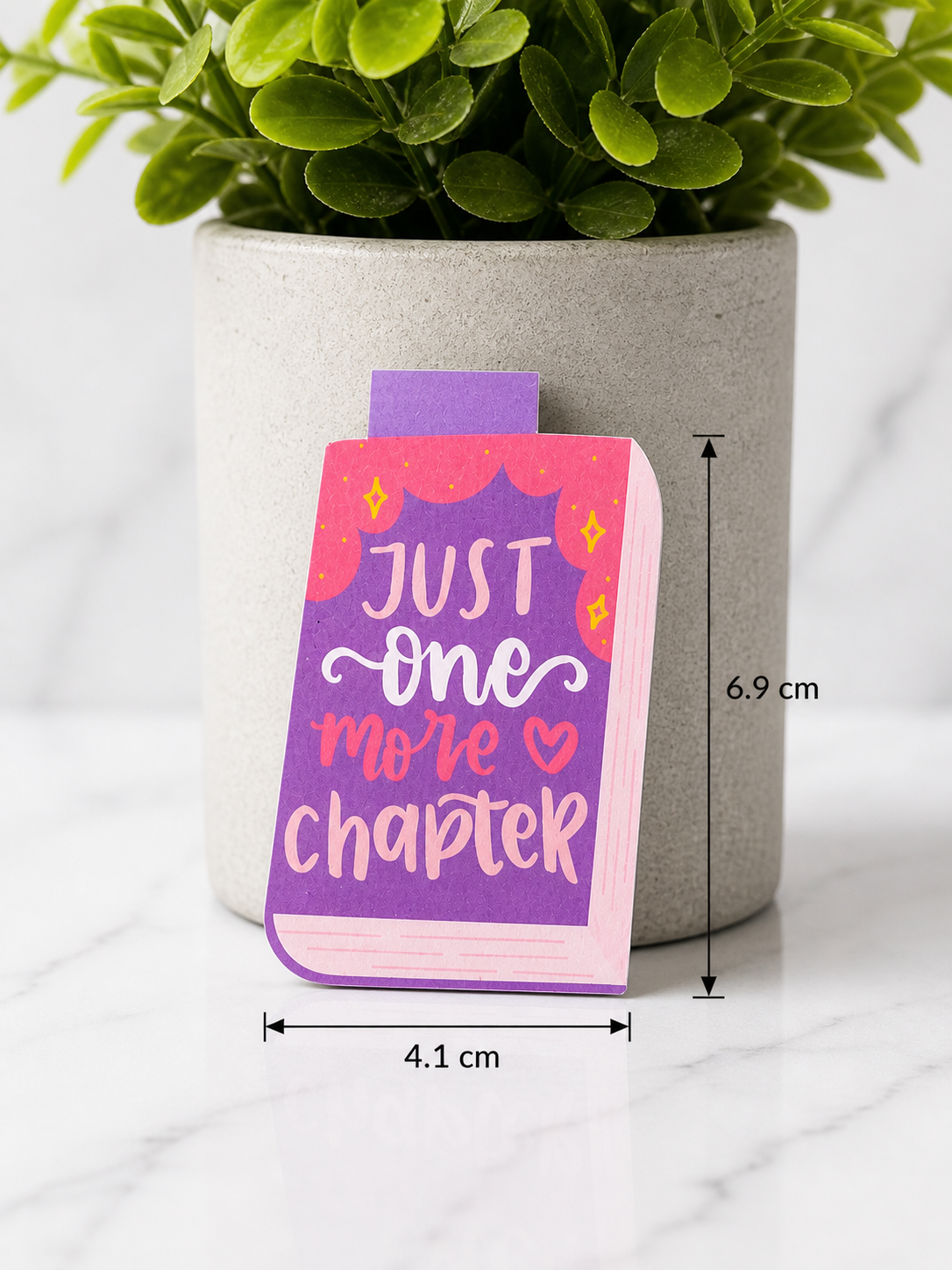 “Just one more chapter” magnetic bookmark with purple and pink book design shown with measurements 6.9 cm by 4.1 cm, placed in front of a plant on a marble surface