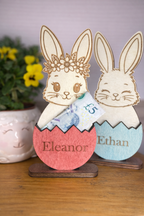 Wooden rabbit money holders in cracked Easter eggs and names 'Eleanor' and 'Ethan' on a wooden stand.