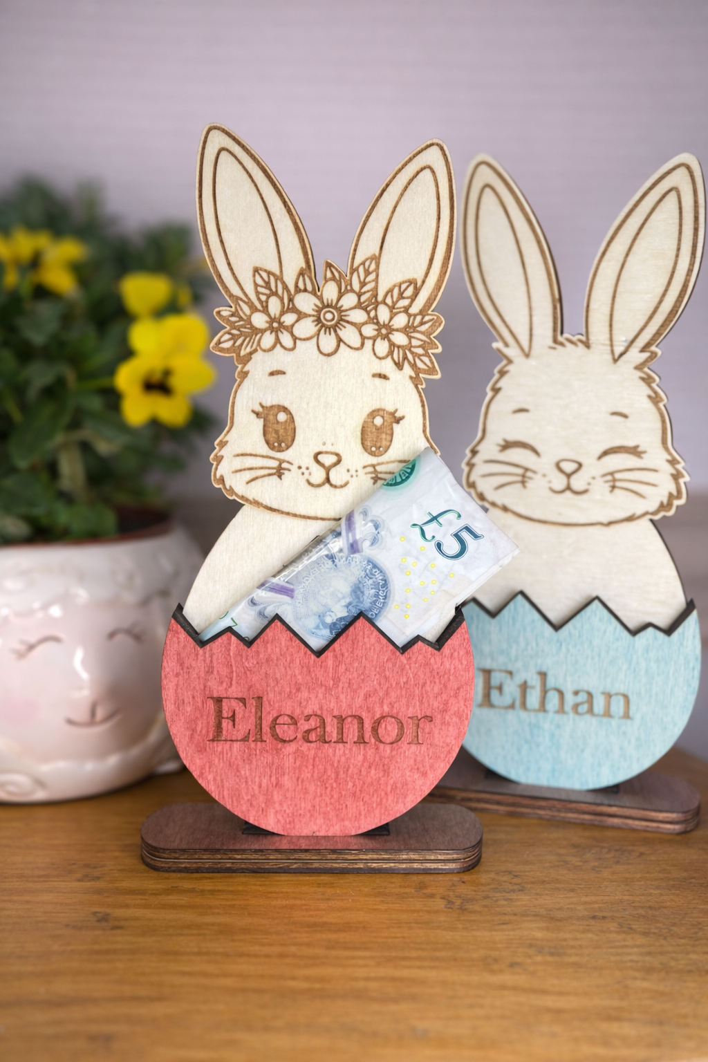 Wooden rabbit money holders in cracked Easter eggs and names 'Eleanor' and 'Ethan' on a wooden stand.