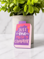 Colourful magnetic bookmark designed as a book with the quote “Just one more chapter” in purple, pink and white, photographed on a marble background with a plant behind