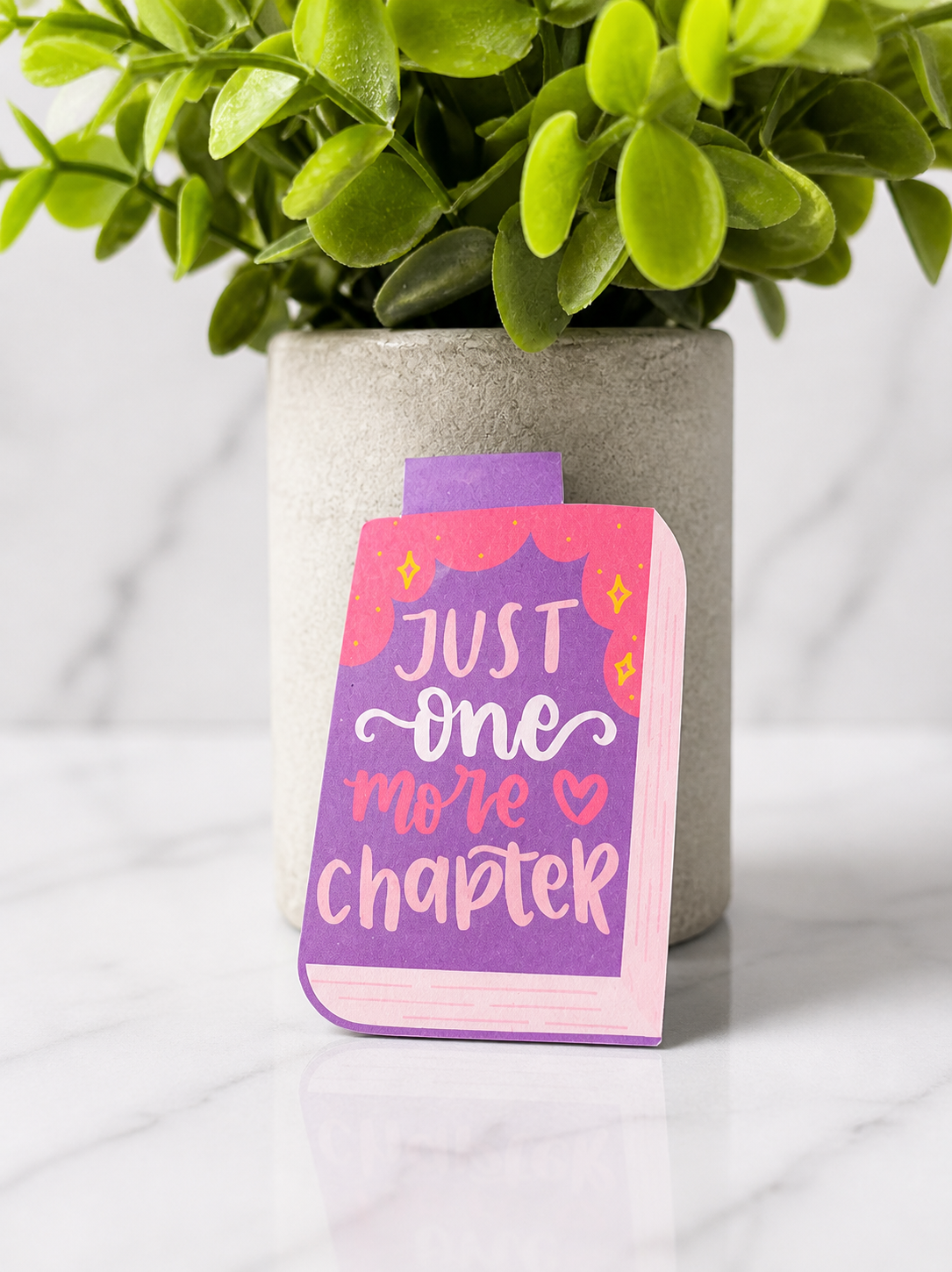 Colourful magnetic bookmark designed as a book with the quote “Just one more chapter” in purple, pink and white, photographed on a marble background with a plant behind