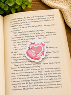 Coquette style pink heart magnetic bookmark resting on an open book with a cosy reading setup including wooden surface, greenery and soft textured fabric