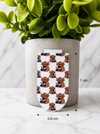 Magnetic bookmark with repeating Highland cow reading book pattern shown with measurements 7 cm by 3.6 cm against a marble background with plant
