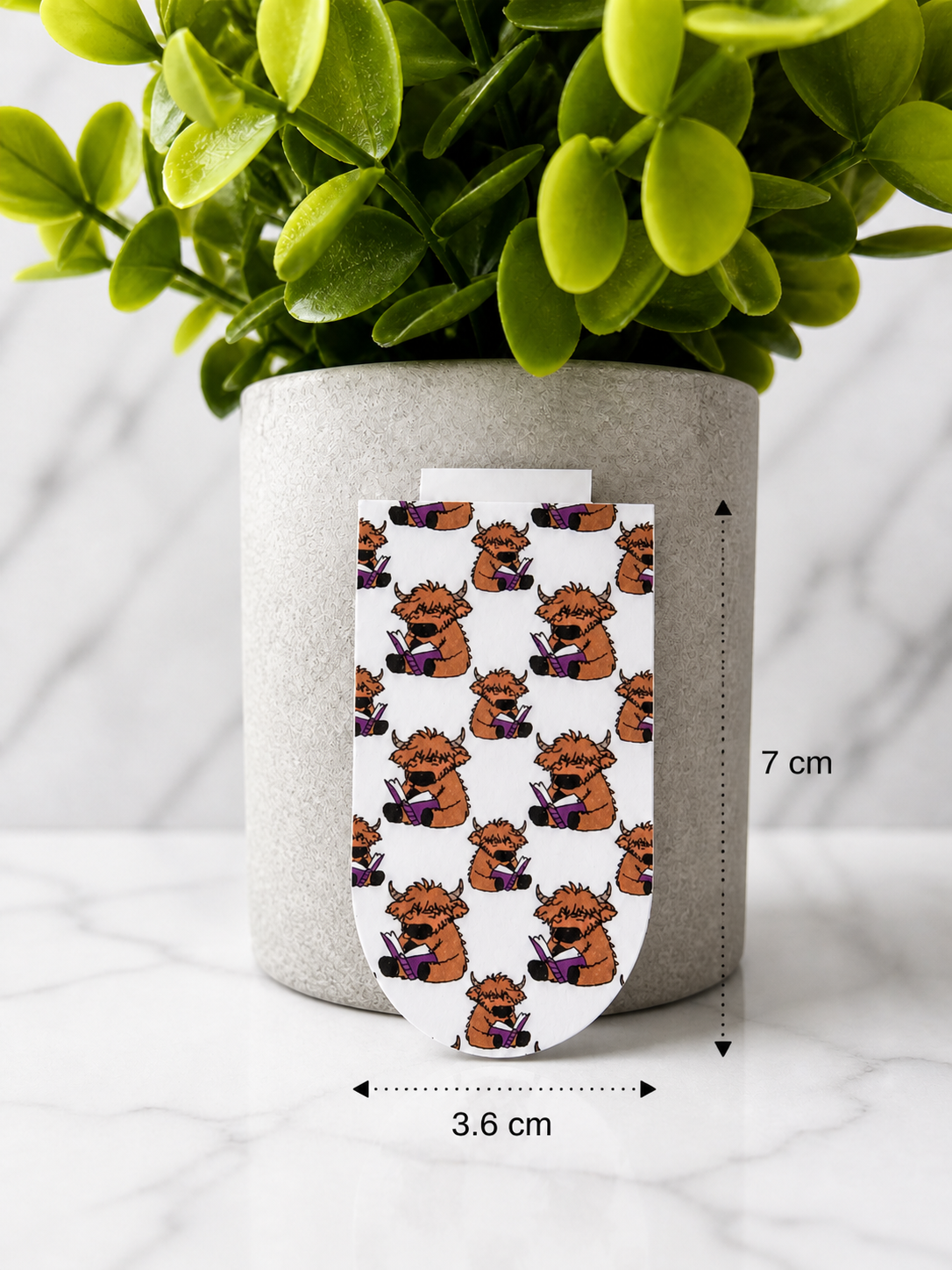 Magnetic bookmark with repeating Highland cow reading book pattern shown with measurements 7 cm by 3.6 cm against a marble background with plant
