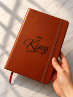 Hand holding personalised brown leather notebook with engraved name and elastic closure on marble background