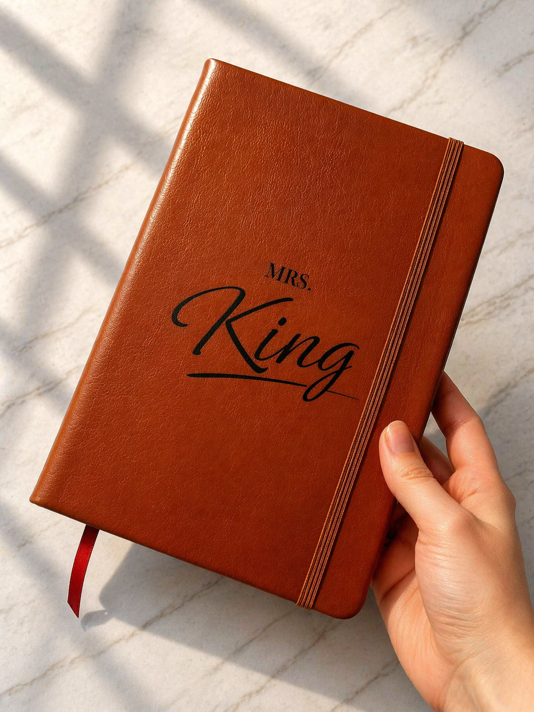 Hand holding personalised brown leather notebook with engraved name and elastic closure on marble background