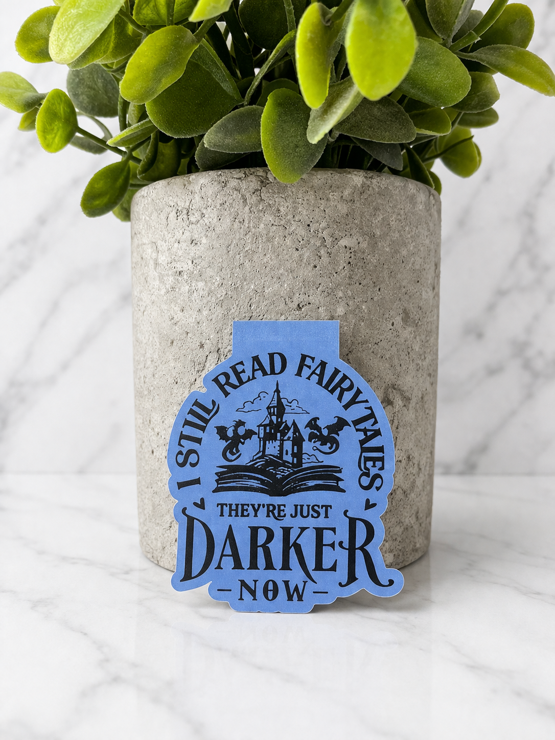 Blue magnetic bookmark with “I still read fairytales they’re just darker now” quote displayed in front of plant