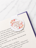 Round floral magnetic bookmark placed at the top of a book page, showing how the magnetic design grips both sides of the page to mark reading progress