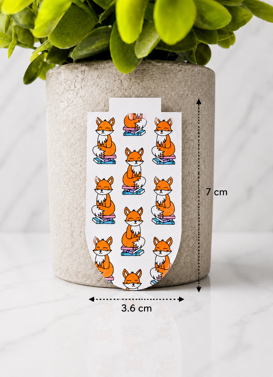 Magnetic bookmark with repeating fox design shown with measurements 7 cm by 3.6 cm on a marble background with plant