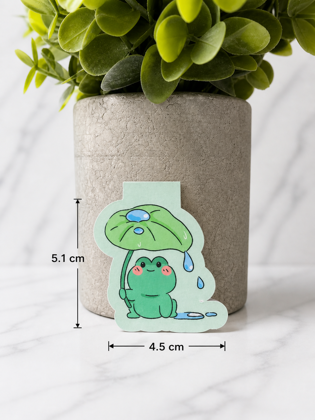 Small cute frog magnetic bookmark with a smiling green frog holding a leaf umbrella, shown against a neutral background with measurements displayed as approximately 5.1 cm tall and 4.5 cm wide
