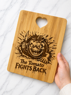 Hand holding bamboo chopping board engraved with angry tomato holding knife and “The Tomato Fights Back” text on marble background