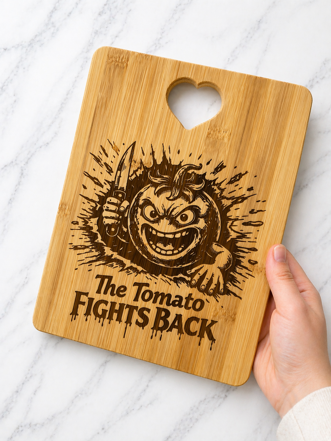 Hand holding bamboo chopping board engraved with angry tomato holding knife and “The Tomato Fights Back” text on marble background