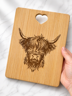 Hand holding engraved bamboo chopping/serving board featuring Highland cow illustration on marble background