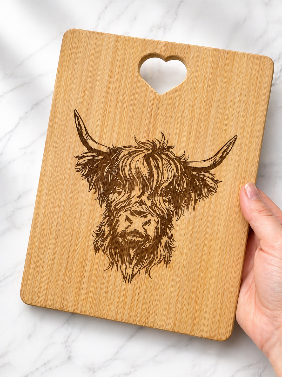 Hand holding engraved bamboo chopping/serving board featuring Highland cow illustration on marble background