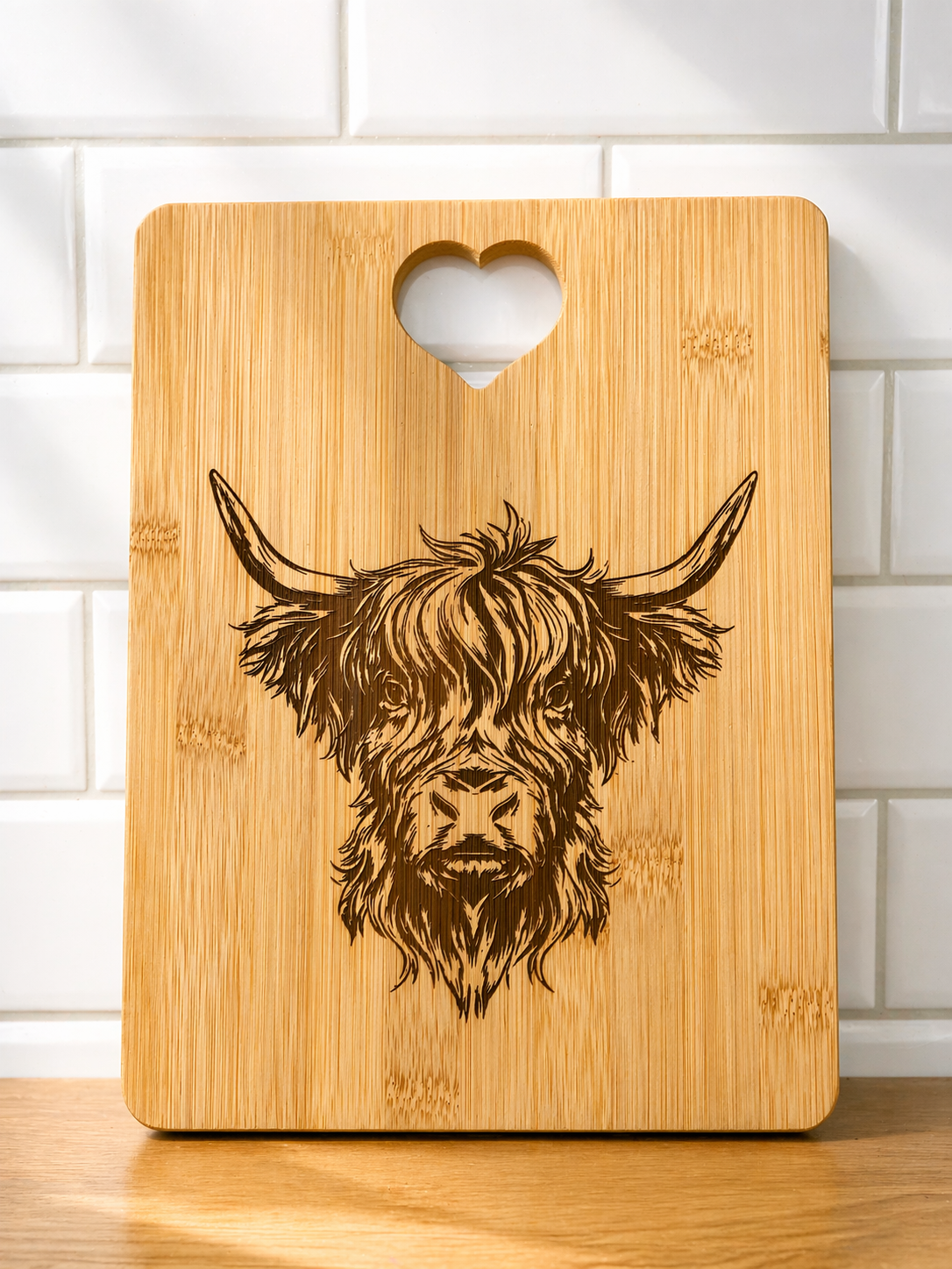 Square bamboo chopping board engraved with detailed Highland cow design and heart handle against tiled kitchen wall