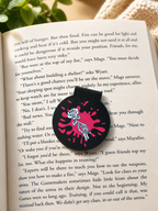 Black gothic bookmark with skeleton hand and rose on open book in warm reading setting