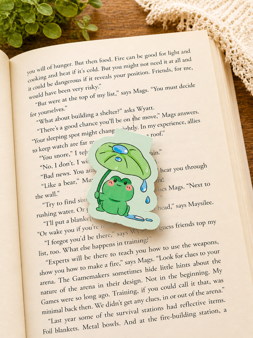 Cute frog magnetic bookmark resting on an open book in a cosy reading setting, with soft lighting and warm textures creating a relaxing aesthetic for book lovers