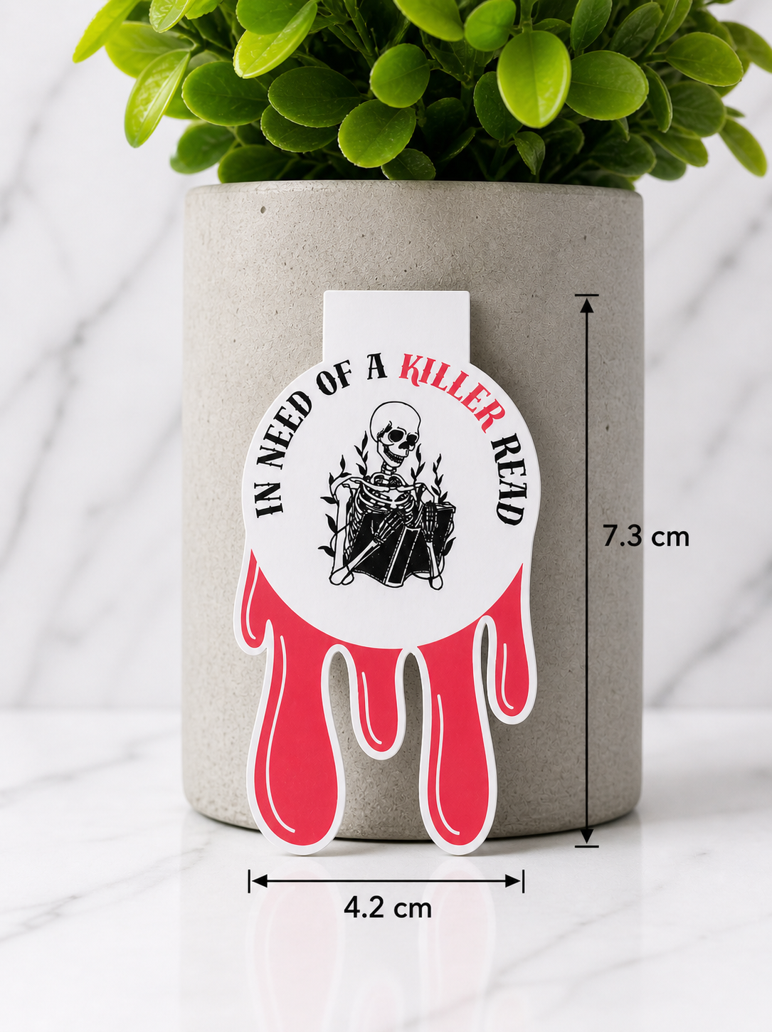 Dripping blood style magnetic bookmark with skeleton reading design and quote “In need of a killer read”, shown with size measurements 7.3 cm by 4.2 cm against a neutral plant and marble background