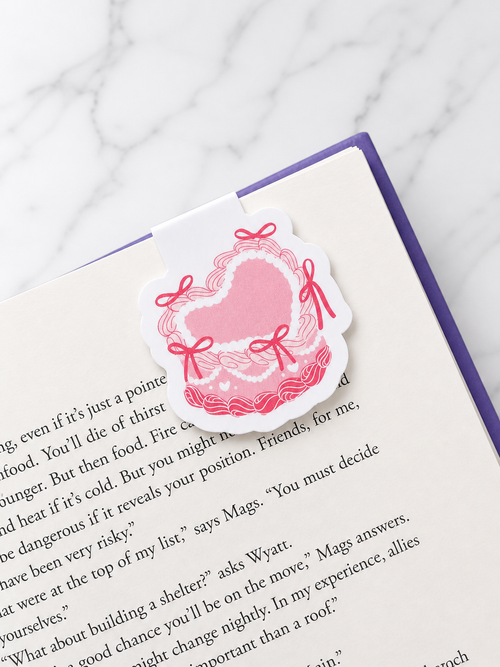 Pink heart magnetic bookmark with bow design clipped onto the top of an open book page, showing how it securely marks pages while reading