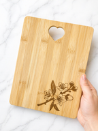 Hand holding a rectangular bamboo chopping board with engraved floral corner design and heart handle on marble background