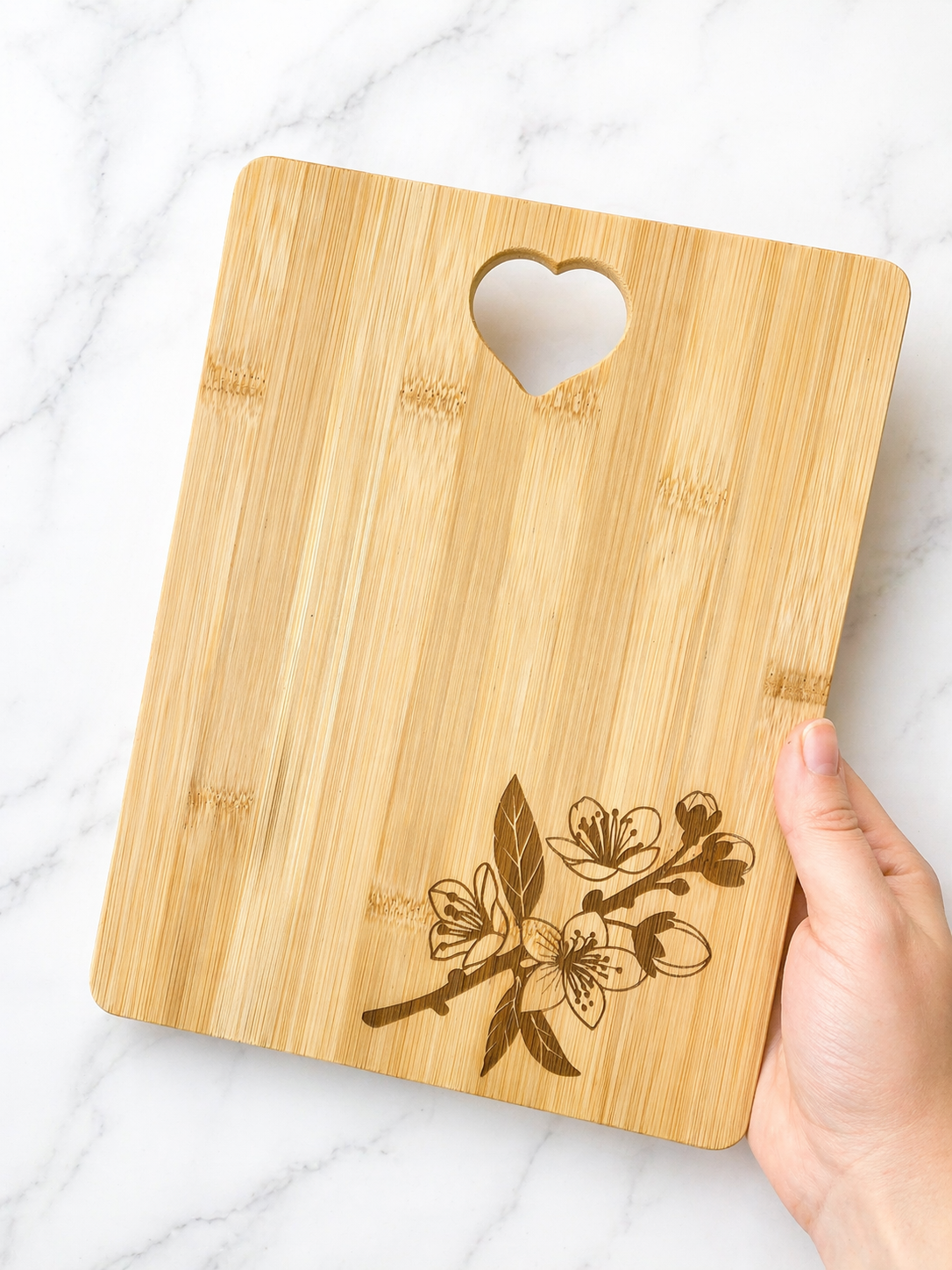 Hand holding a rectangular bamboo chopping board with engraved floral corner design and heart handle on marble background