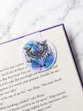 Round magnetic bookmark with moth and crystal design clipped onto a book page
