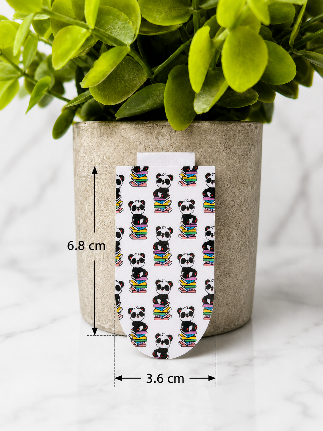 Magnetic bookmark with repeating panda sitting on books pattern shown with measurements 6.8 cm by 3.6 cm on a marble background with plant