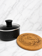 Engraved cork trivet with floral wreath design next to cooking pot on marble kitchen surface