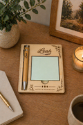 Personalized woden sticky note holder with pen on a wooden desk with a plant, framed picture, and candle.