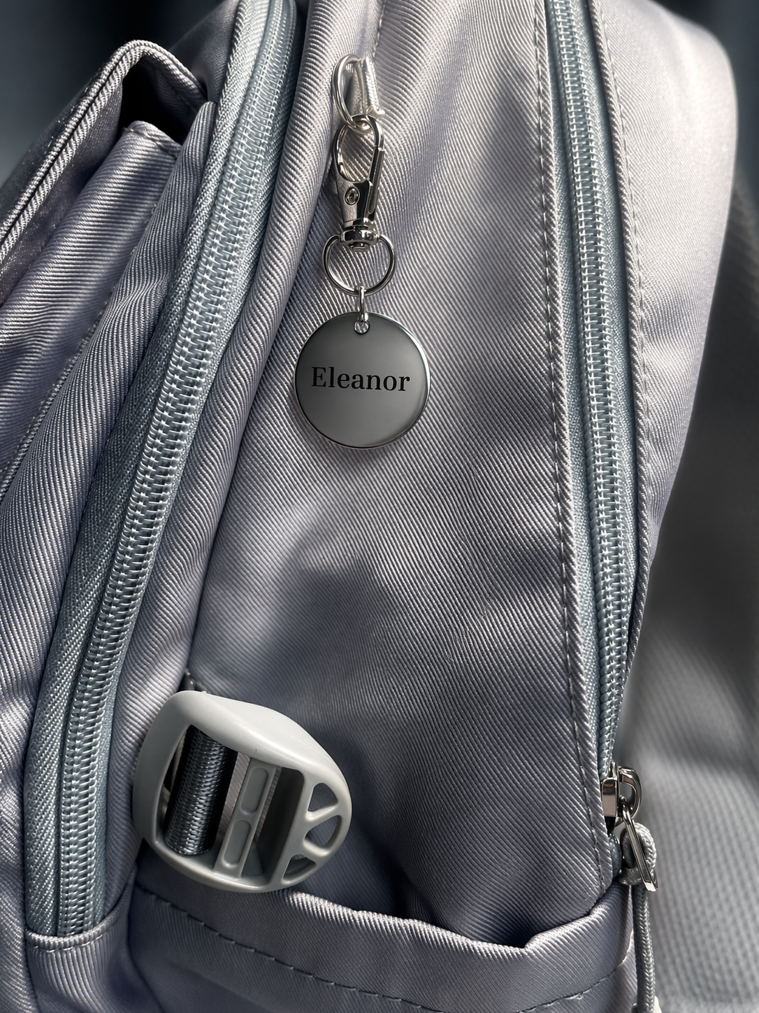 Personalised metal keychain with name Eleanor attached to grey backpack zip, custom engraved bag tag