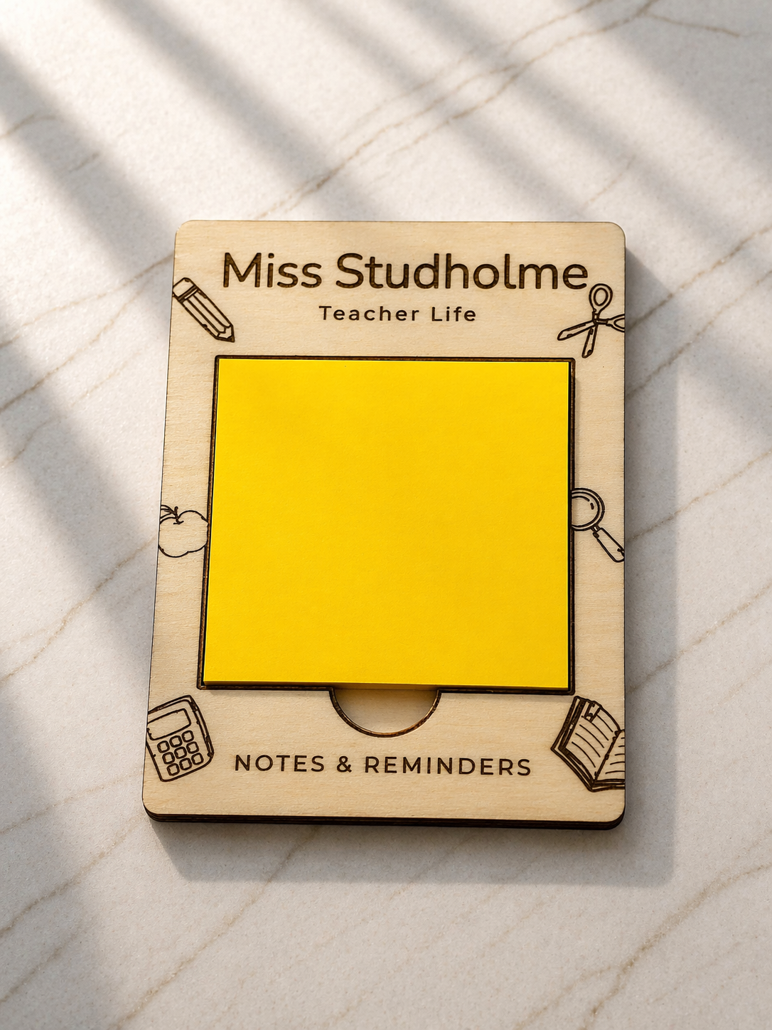 Close-up of engraved wooden sticky note holder personalised with teacher name, different engraved designs such as a book, calculator, pencil and more. Sticky notes on marble surface
