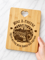 Hand holding engraved bamboo wine and cheese board with vineyard design and heart cut-out handle on marble background
