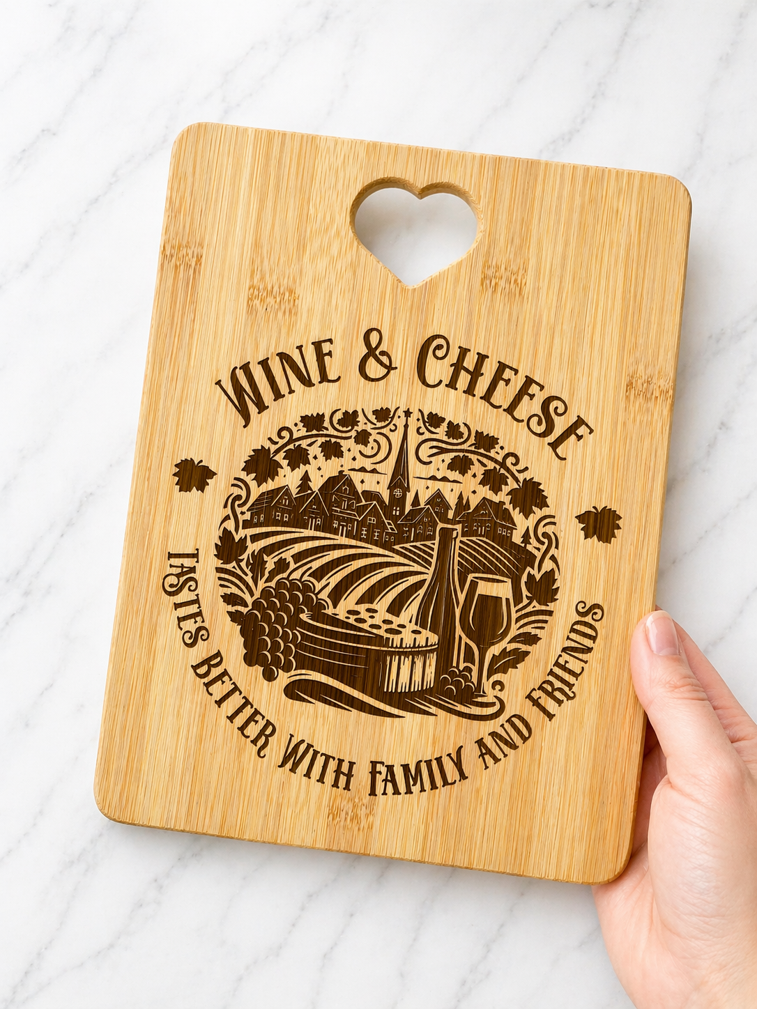 Hand holding engraved bamboo wine and cheese board with vineyard design and heart cut-out handle on marble background