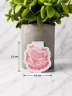 Pink coquette heart magnetic bookmark with bow details shown with size measurements 5.2 cm by 4.5 cm, displayed in front of a neutral plant pot on a marble background