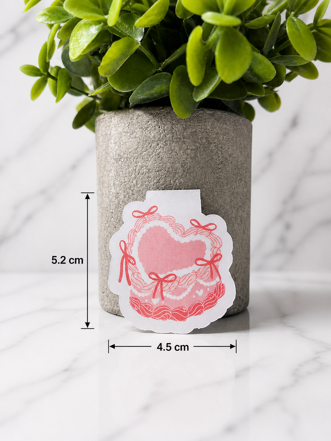 Pink coquette heart magnetic bookmark with bow details shown with size measurements 5.2 cm by 4.5 cm, displayed in front of a neutral plant pot on a marble background