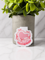 Cute pink heart magnetic bookmark with delicate bows and lace-style illustration, standing upright on a white marble surface with a decorative plant in the background