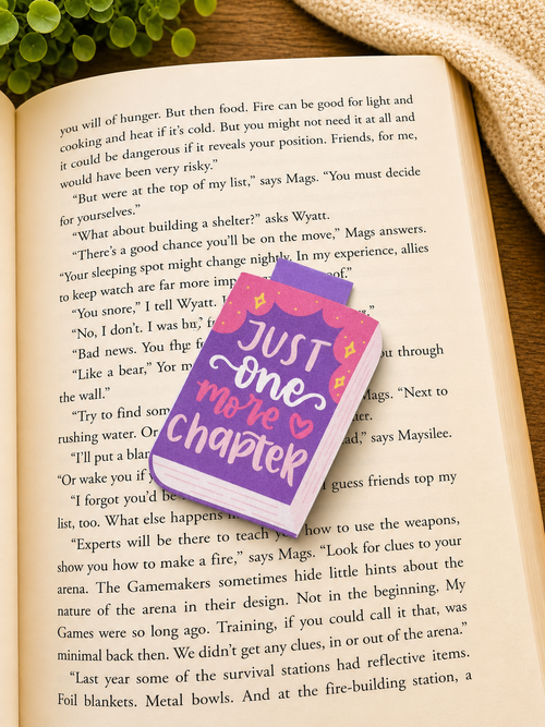“Just one more chapter” magnetic bookmark resting on an open book in a cosy reading setting with warm lighting, greenery and textured fabric background