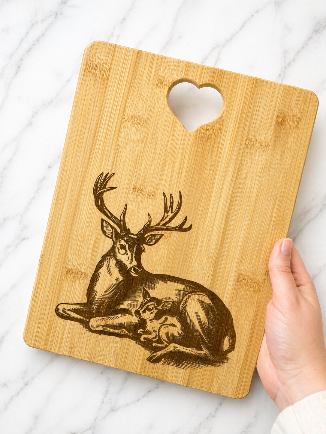 Hand holding bamboo chopping board with detailed deer and fawn engraving and heart handle on marble background