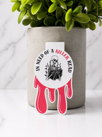 Dark romance magnetic bookmark featuring a skeleton reading a book with red dripping blood design and the phrase “In need of a killer read”, displayed on a white marble surface with plant background