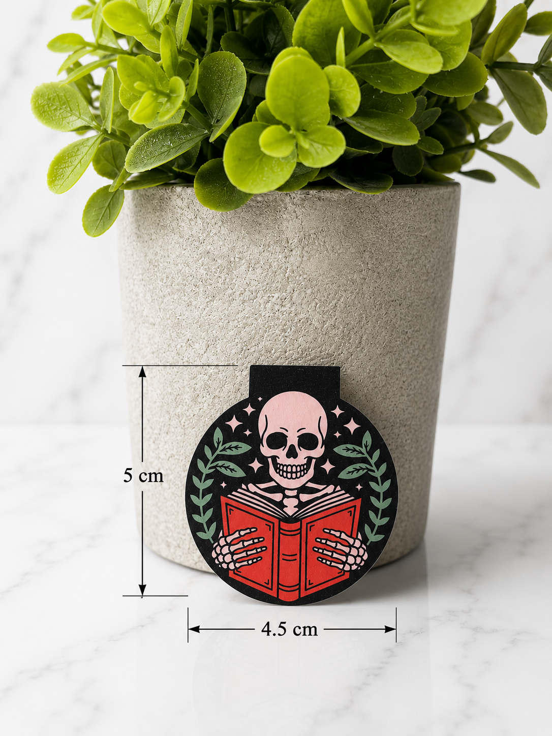 Magnetic bookmark featuring a skeleton reading a red book shown with measurements 5 cm by 4.5 cm against a marble background with plant