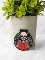 Magnetic bookmark with skeleton reading a red book surrounded by leaves and stars on a neutral background