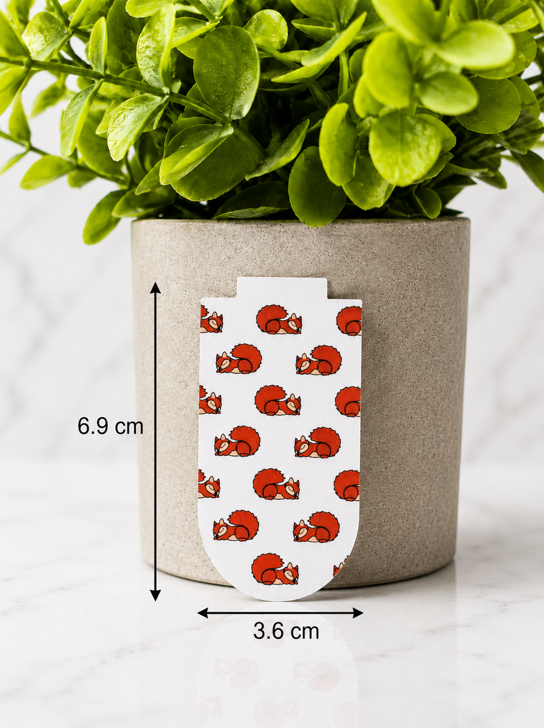 Magnetic bookmark with sleeping squirrel pattern shown with measurements 6.9 cm by 3.6 cm on a marble background with plant