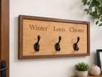 Wooden wall hook with three hooks and engraved names on a white wall.