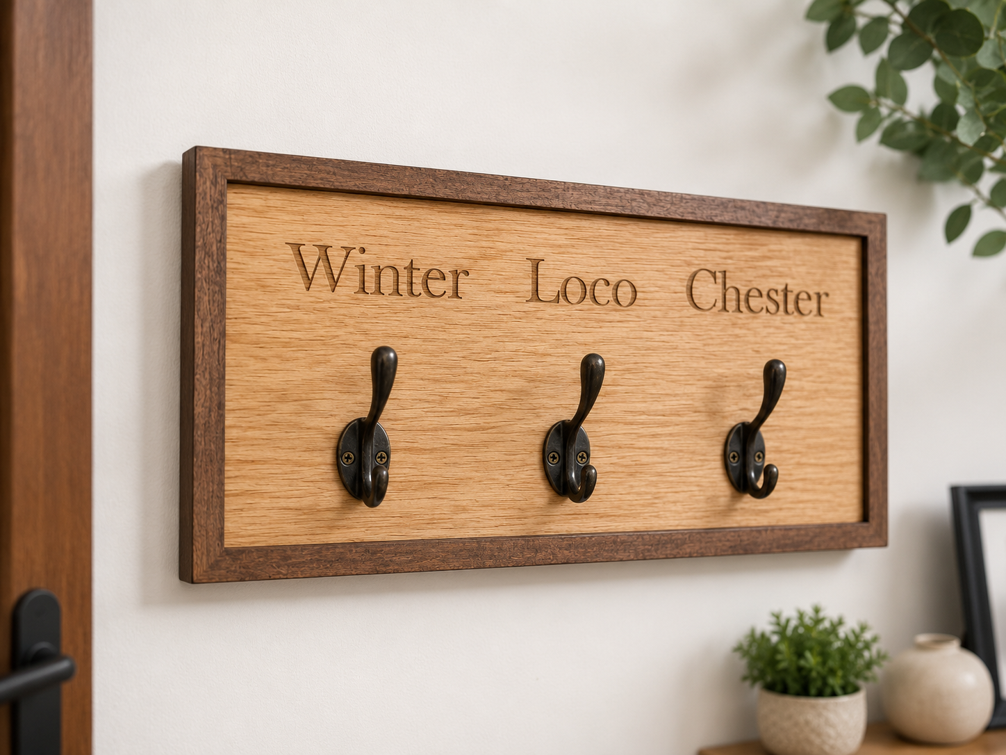 Wooden wall hook with three hooks and engraved names on a white wall.