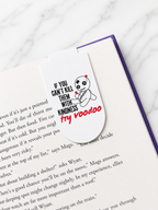 Magnetic bookmark clipped onto the top of an open book page featuring a voodoo doll design and humorous text, demonstrating how it securely marks pages